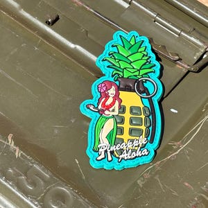 May include: A colorful patch featuring a cartoon pineapple grenade design. The grenade is yellow with a green top and a blue outline. A cartoon woman in a hula skirt is on the side, with the words "Pineapple Aloha".