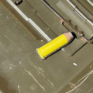 May include: A yellow and brown bullet rests on a green metal surface. The bullet has a yellow casing and a brown tip. The surface appears to be part of a container, with hinges and a handle visible.