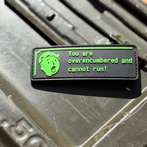 May include: A black and green patch with the text "You are overencumbered and cannot run!" and a green cartoon character. The patch is rectangular and has a green border. The background is a textured, olive green surface.