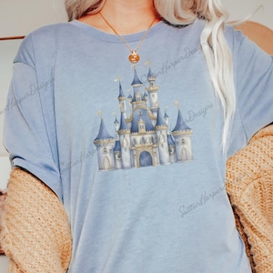 May include: A light blue t-shirt with a watercolor illustration of a fairytale castle. The castle is blue, gold, and white with a pointed roof and towers. The t-shirt has the text "Sutton Harper Designs" printed on the bottom right corner.