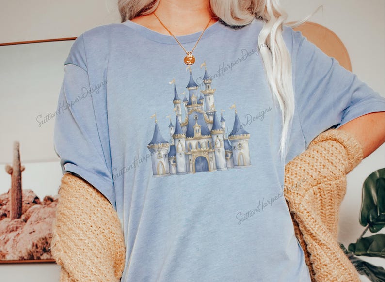 May include: A light blue t-shirt with a watercolor illustration of a fairytale castle. The castle is blue, gold, and white with a pointed roof and towers. The t-shirt has the text "Sutton Harper Designs" printed on the bottom right corner.