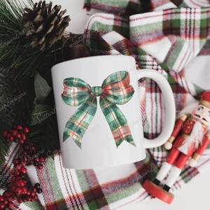 Puede incluir: Taza de cer&aacute;mica blanca con un dise&ntilde;o de lazo de cuadros verdes y rojos. La taza est&aacute; sobre una tela a cuadros con una figura de cascanueces.