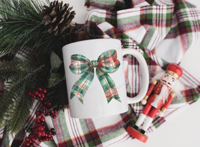 Puede incluir: Taza de cer&aacute;mica blanca con un dise&ntilde;o de lazo de cuadros verdes y rojos. La taza est&aacute; sobre una tela a cuadros con una figura de cascanueces.
