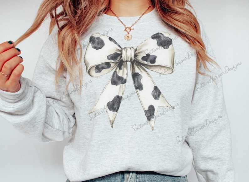 Puede incluir: Una sudadera gris con un gran lazo de estampado de vaca blanco y negro en la parte delantera. El lazo est&aacute; atado en un nudo y los extremos caen por la parte delantera de la camiseta.