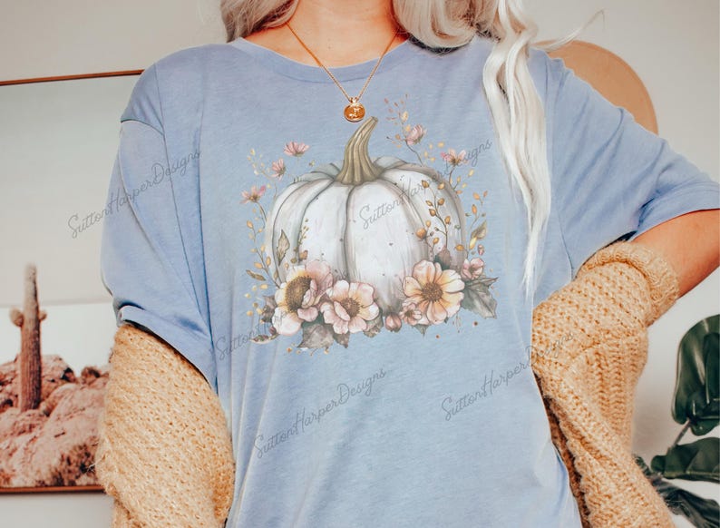 May include: A light blue t-shirt with a white pumpkin surrounded by pink and brown flowers. The text "Sutter Harper Designs" is printed on the shirt.