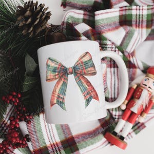May include: White ceramic mug featuring a red, green and white plaid bow design. The mug is resting on a plaid fabric with a nutcracker figurine.