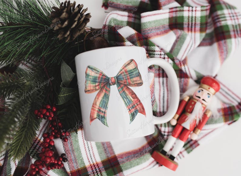 May include: White ceramic mug featuring a red, green and white plaid bow design. The mug is resting on a plaid fabric with a nutcracker figurine.