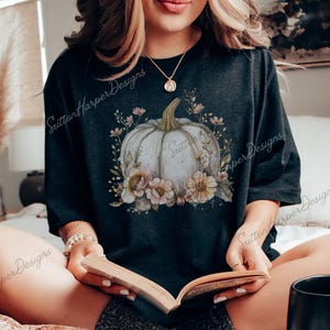 May include: A black t-shirt with a white pumpkin and floral design. The pumpkin is surrounded by pink and yellow flowers and leaves. The text "Sutton Harper Designs" is visible on the shirt.