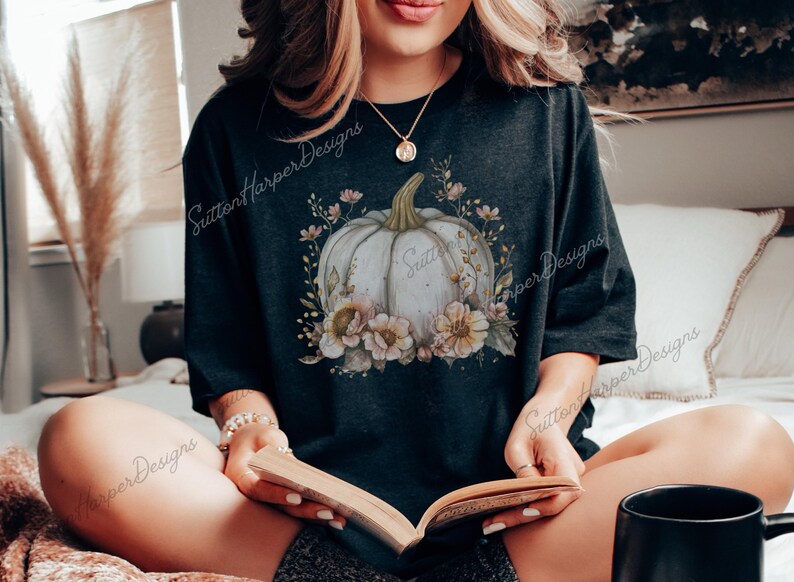 May include: A black t-shirt with a white pumpkin and floral design. The pumpkin is surrounded by pink and yellow flowers and leaves. The text "Sutton Harper Designs" is visible on the shirt.