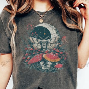 Puede incluir: Una camiseta gris oscuro con un estampado gr&aacute;fico de una polilla, setas y una luna creciente. La polilla es blanca con marcas negras y tiene las alas extendidas. Las setas son rojas con manchas blancas. La luna creciente es blanca con un contorno negro.