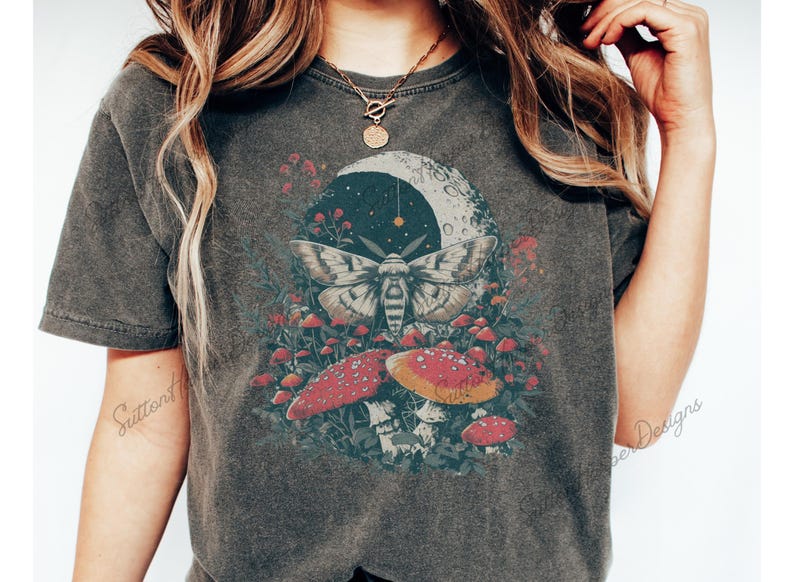 Puede incluir: Una camiseta gris oscuro con un estampado gr&aacute;fico de una polilla, setas y una luna creciente. La polilla es blanca con marcas negras y tiene las alas extendidas. Las setas son rojas con manchas blancas. La luna creciente es blanca con un contorno negro.