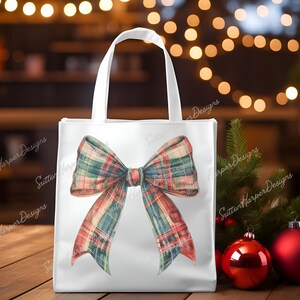 May include: White tote bag featuring a large red, green and white plaid bow. The bow is tied in a knot with the ends hanging down. The bag is placed on a wooden surface.