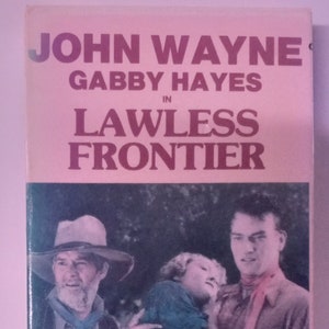 May include: A vintage movie poster featuring John Wayne and Gabby Hayes in the film "Lawless Frontier". The poster shows a scene with John Wayne holding a young girl in his arms, while Gabby Hayes stands beside him.