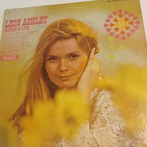 May include: A vintage vinyl record cover featuring a woman with blonde hair and a white lace top. The cover is yellow and features a pink flower with hearts. The text on the cover reads "Leon Ashley Flower of Love".