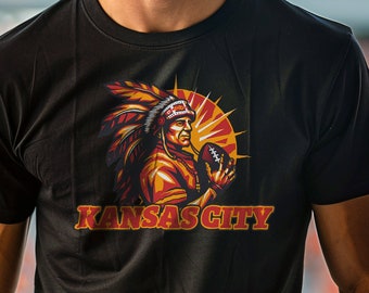 May include: Black t-shirt featuring a Native American chief holding a football, with the text "KANSAS CITY" in red and gold. The design includes a sunburst background. The shirt is worn by a person.