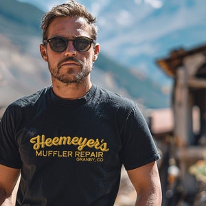 May include: A man wearing sunglasses and a black t-shirt with the text "Heemeyers Muffler Repair Granby, CO" in yellow. He is standing in front of a mountain range.