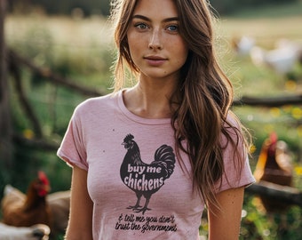 May include: A woman with long brown hair and freckles is wearing a pink t-shirt with a black and white illustration of a chicken and the text "buy me chickens & tell me you don't trust the government".