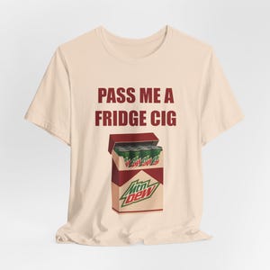 以下が含まれることがあります： ベージュ色のTシャツ。マルーン色の文字で「PASS ME A FRIDGE CIG」と書かれています。Tシャツには、マウンテンデューの缶が入ったタバコの箱のグラフィックが描かれています。箱は赤と白で、マウンテンデューのロゴが入っています。