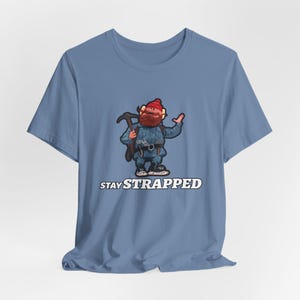 Könnte beinhalten: Hellblaues T-Shirt mit einer Cartoon-Illustration eines bärtigen Bergsteigers mit einer Spitzhacke. Der Text "STAY STRAPPED" ist darunter in Weiß gedruckt.