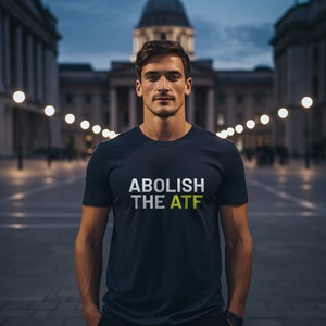 Può includere: Un uomo indossa una maglietta blu scuro con la scritta "ABOLISH THE ATF" stampata in lettere bianche e gialle. È in piedi davanti a un edificio con le luci.
