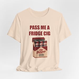 May include: A light beige t-shirt with the text "PASS ME A FRIDGE CIG" in maroon. Below the text is a graphic of a Dr. Pepper Zero Sugar carton with four cans. The shirt is a casual, short-sleeved style.