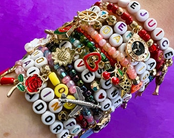 Peut inclure: Plusieurs bracelets de perles colorées avec des breloques variées, dont une rose, un avion et un cœur. Certains bracelets comportent des perles blanches avec des lettres épelant des mots comme "ADORE" et "LOVE". Fond violet.