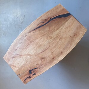 Könnte beinhalten: Ein hellfarbener Holztisch mit einer gebogenen Kante. Die Tischplatte weist natürliche Holzmaserungsmuster und eine dunkle, lineare Intarsie auf. Der Tisch ist rechteckig mit abgerundeten Ecken und zeigt ein rustikales Design.