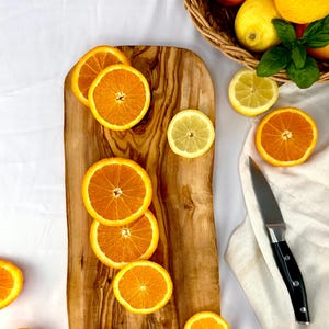 May include: A wooden chopping board with orange and lemon slices. A black-handled knife rests on a white cloth. A basket of lemons and oranges is in the background. Fresh fruit and kitchenware.