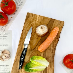 May include: A wooden chopping board with a stainless steel knife, a carrot, a mushroom, and lettuce. Two red tomatoes and a garlic bulb are also visible. The board has a natural wood grain pattern.
