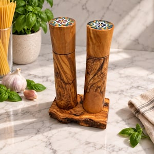 Könnte beinhalten: Zwei zylindrische Salz- und Pfeffermühlen aus Holz mit natürlicher Maserung und bunten, blumenverzierten Keramikdeckeln, die auf einem passenden rechteckigen Holzsockel mit einer rauen, organischen Kante stehen.