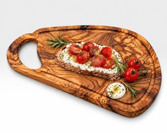 Op de afbeelding: Een onregelmatig, ovaalvormig houten serveerplank met een opvallende houtnerf en een ronde handgreep. Een snee geroosterd brood belegd met witte kaas, gehalveerde rode cherrytomaatjes en groene kruiden ligt op de plank.