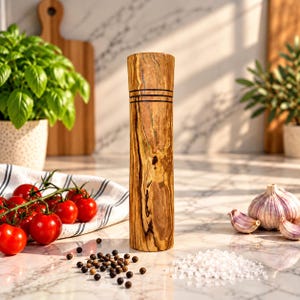 Könnte beinhalten: Eine hohe, zylindrische Pfeffermühle aus Holz mit natürlicher hellbrauner Maserung, dunkelbraunen Streifen und zwei dünnen, horizontalen schwarzen Ringen am oberen Ende. Die Mühle steht aufrecht auf einer Marmoroberfläche mit Pfefferkörnern.