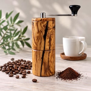 Könnte beinhalten: Eine zylindrische manuelle Kaffeemühle aus Olivenholz mit ausgeprägter dunkler Maserung. Sie verfügt über einen silbernen Metallgriff mit einem schwarzen Knauf. Daneben liegen verstreute Kaffeebohnen und ein Häufchen Kaffeepulver.