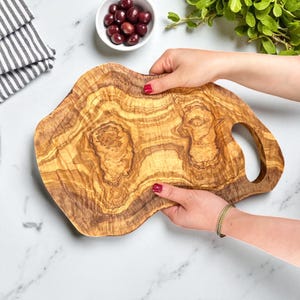 May include: An olive wood chopping board with a distinctive, organic form and a built-in handle. The board displays a rich, warm colour with intricate wood grain patterns. The board is held by a person with red nail polish. The board measures approximately 38 cm long.