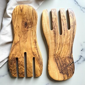Peut inclure: Deux couverts à salade en bois d'olivier. L'un a une large poignée et trois dents, l'autre a un design similaire. Le bois présente un veinage naturel. Idéal pour servir des salades. Les couverts sont en bois.