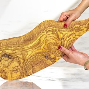 May include: A large, uniquely shaped wooden cutting board with a natural olive wood grain pattern. The board is held by two hands with red nail polish, showcasing its organic form and warm tones. The board is approximately 50 cm long.