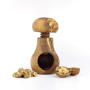 Op de afbeelding: Een houten notenkraker met een paddenstoelvormige bovenkant en een schroefmechanisme. De notenkraker is gemaakt van lichtbruin hout en heeft een ronde opening. Walnoten en gepelde walnoten liggen naast de notenkraker.