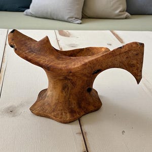 Könnte beinhalten: Eine handgefertigte Holzschale mit einer einzigartigen, organischen Form. Die Schale besteht aus hellbraunem Holz mit einem ausgeprägten Maserungsmuster und dunklen Knoten. Das Design weist einen ausgestellten Rand und einen stabilen Sockel auf, geeignet für dekorative Zwecke.