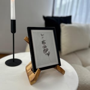 May include: A black framed print of a botanical illustration, displayed on a wooden easel. A lit white candle sits in a black candlestick holder. The items are on a white table.