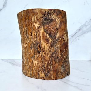 Könnte beinhalten: Zylindrisches Holzobjekt mit rauer, strukturierter Oberfläche. Das Holz hat eine natürliche, warme braune Farbe mit dunkleren Streifen und Mustern. Das Objekt scheint ein Abschnitt eines Baumstamms zu sein, möglicherweise für dekorative oder funktionale Zwecke.