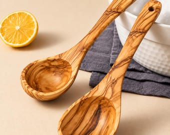 Może przedstawiać: Dwie drewniane łyżki wykonane z drewna oliwnego, charakteryzujące się wyraźnym naturalnym usłojeniem w odcieniach jasnego i ciemnego brązu. Każda łyżka ma głęboką, zaokrągloną miseczkę oraz uchwyt z małym otworem do zawieszenia.