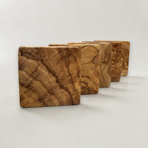 Könnte beinhalten: Ein Set aus vier quadratischen Untersetzern aus hellbraunem Holz mit einer markanten, wirbelnden dunkelbraunen Maserung. Jeder Untersetzer hat eine glatte, flache Oberfläche und abgerundete Kanten und ist in einer versetzten Reihe angeordnet.