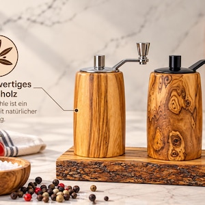 K&ouml;nnte beinhalten: Zwei zylindrische Gew&uuml;rzm&uuml;hlen aus Olivenholz mit Metallkurbeln, eine mit silberfarbenem und eine mit schwarzem Aufsatz. Sie stehen auf einem rechteckigen Olivenholzbrett mit nat&uuml;rlicher Baumkante und ausgepr&auml;gter Maserung.