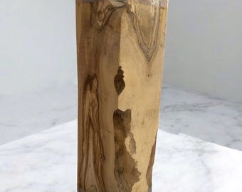 May include: A tall, rectangular wooden sculpture with a natural, light brown finish. The wood grain is visible, with darker brown patterns. The top has a darker, polished appearance. The sculpture stands on a white marble surface.