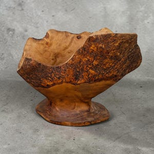 May include: A natural-edged wooden bowl crafted from a single piece of wood, featuring a prominent, rough, dark brown bark exterior and a smooth, polished, light golden-brown interior grain. The bowl rests on a wide, flat, circular wooden base.
