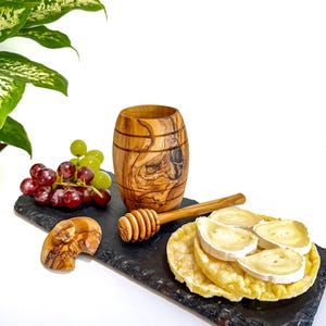 Puede incluir: Un recipiente de madera en forma de barril, un cucharón de miel y una pequeña escultura de madera están dispuestos sobre una tabla negra con uvas y galletas saladas cubiertas de queso. Los elementos están sobre un fondo blanco.