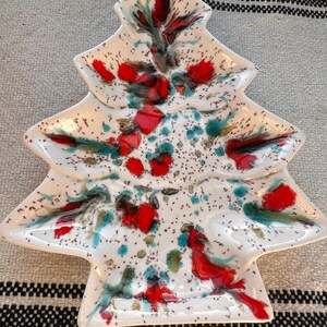 May include: A white ceramic Christmas tree-shaped serving dish with a speckled pattern. The dish is decorated with red, teal, and green abstract designs. The dish is on a neutral-colored striped surface.
