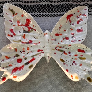 May include: A white ceramic butterfly-shaped dish with red, orange, and black splatter paint details. The dish has a glossy finish and is designed with wing-shaped compartments. The butterfly dish is a decorative item.