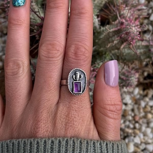 May include: A silver ring featuring a purple gemstone set within a detailed design resembling a cactus. The ring is worn on a finger with light blue and green glitter nail polish.