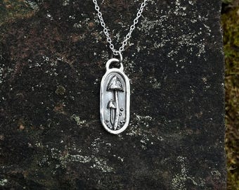 Puede incluir: Un collar con colgante de plata con un diseño de seta. El colgante tiene forma ovalada con un borde texturizado y una pequeña anilla. La cadena es de plata fina. El collar se muestra sobre una piedra oscura y texturizada. El colgante mide unos 2,5 cm de largo.